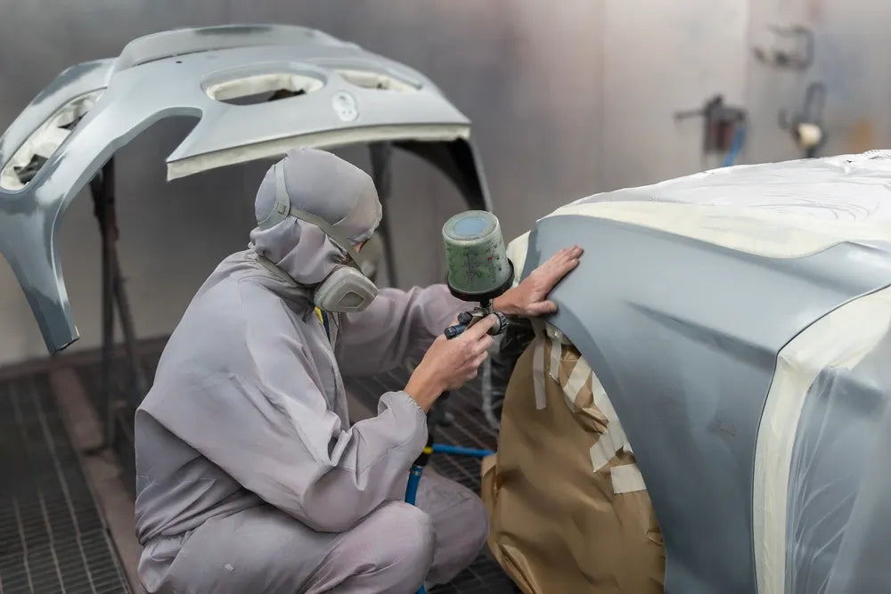 Worker wearing a respirator and protective suit uses a spray gun to paint a primed car surface in a professional auto body workshop.
