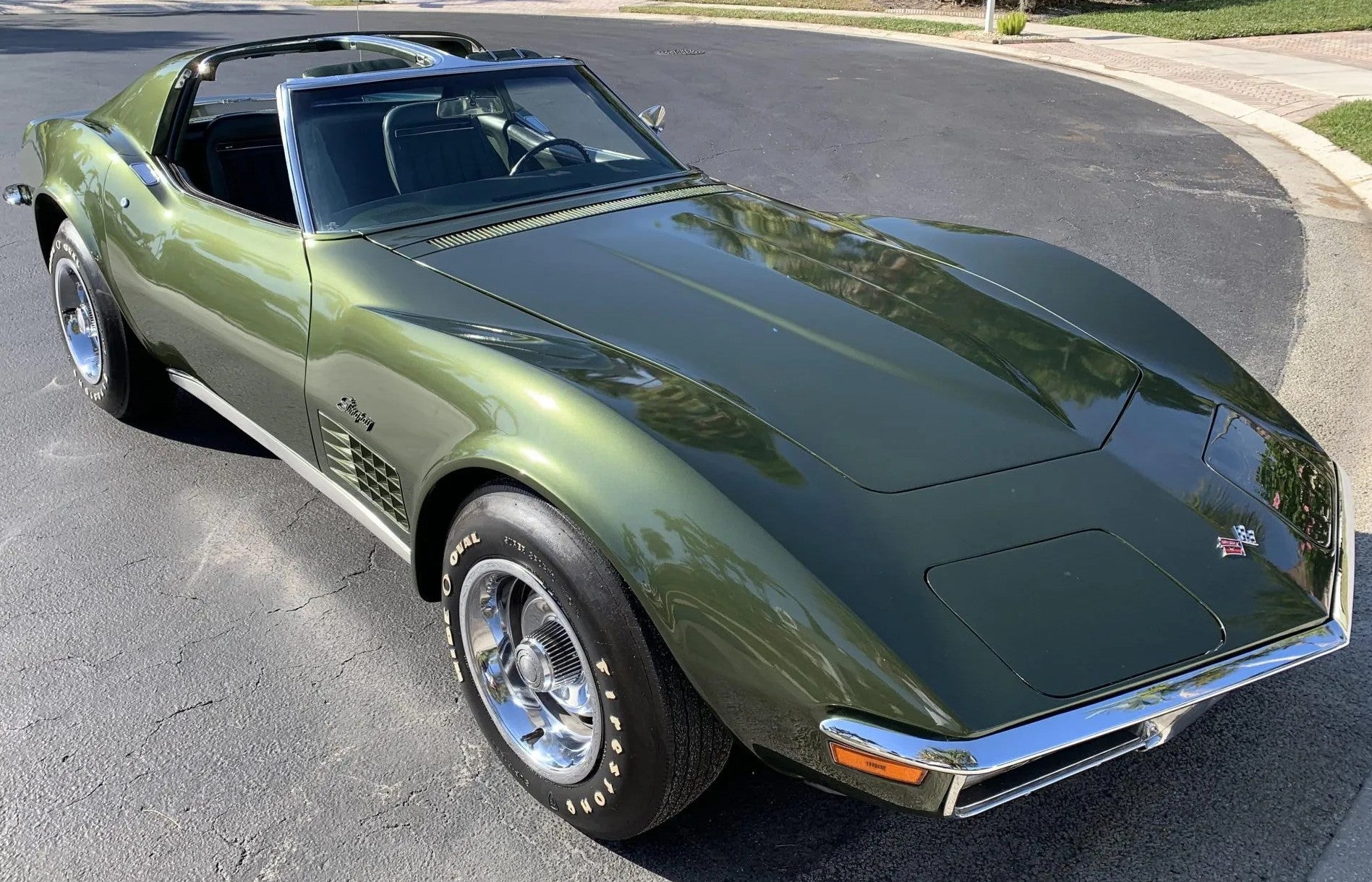 A classic olive green convertible sports car parked on an asphalt street, showing a three-quarter front view with chrome wheels and a black soft top.